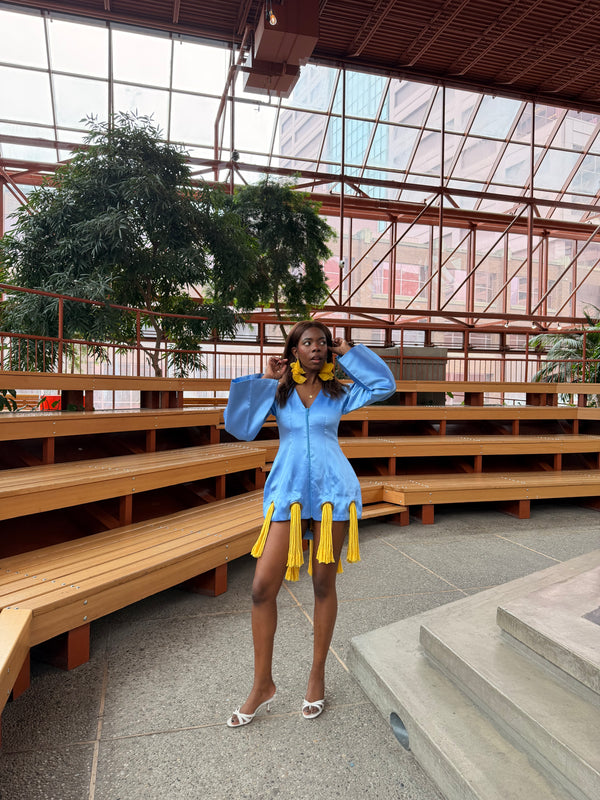 Woman in a blue dress with yellow accents standing in an indoor setting with large windows and wooden benches.