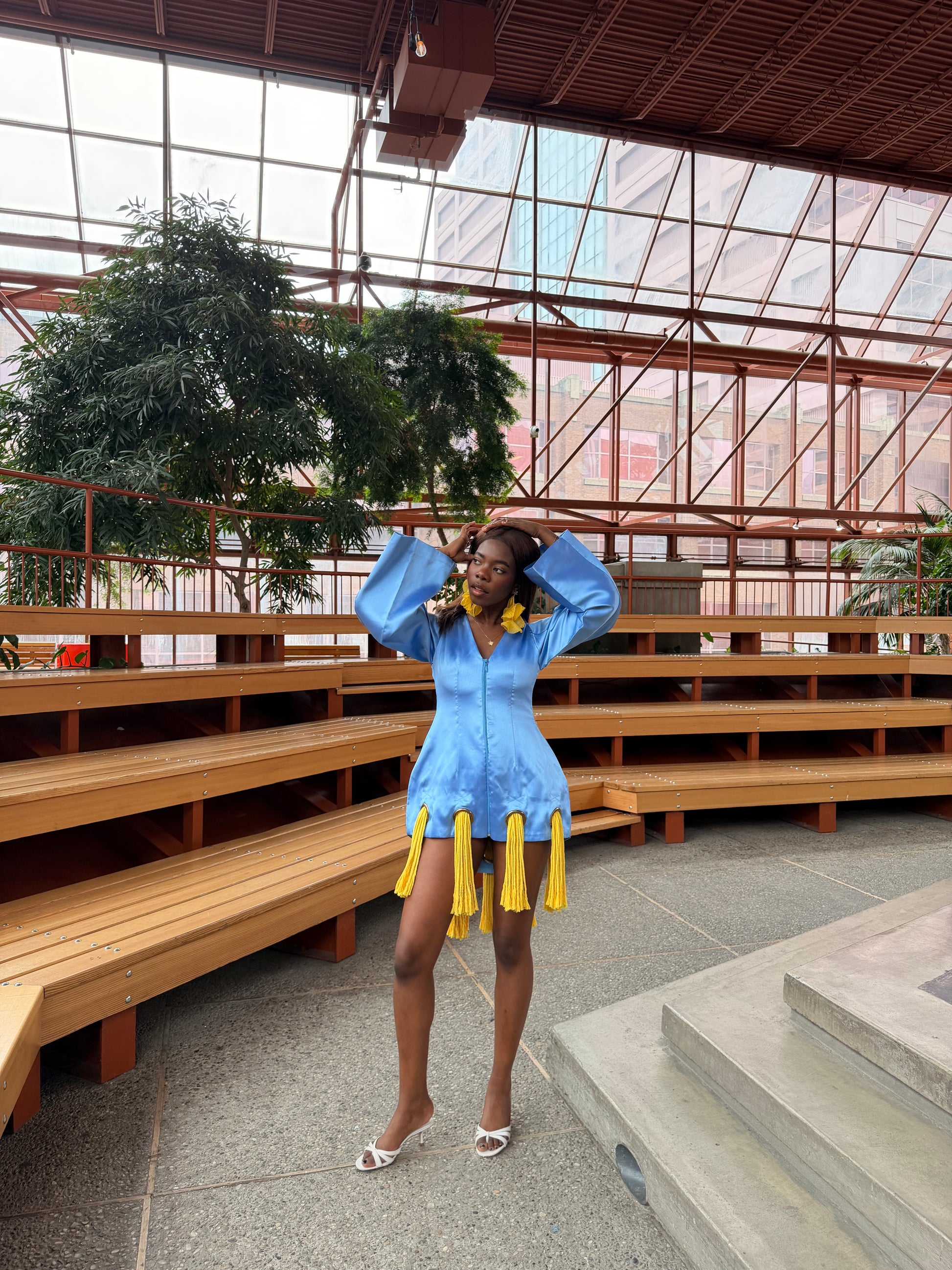 Person in a blue dress with yellow details posing in a large indoor space with wooden benches and plants.